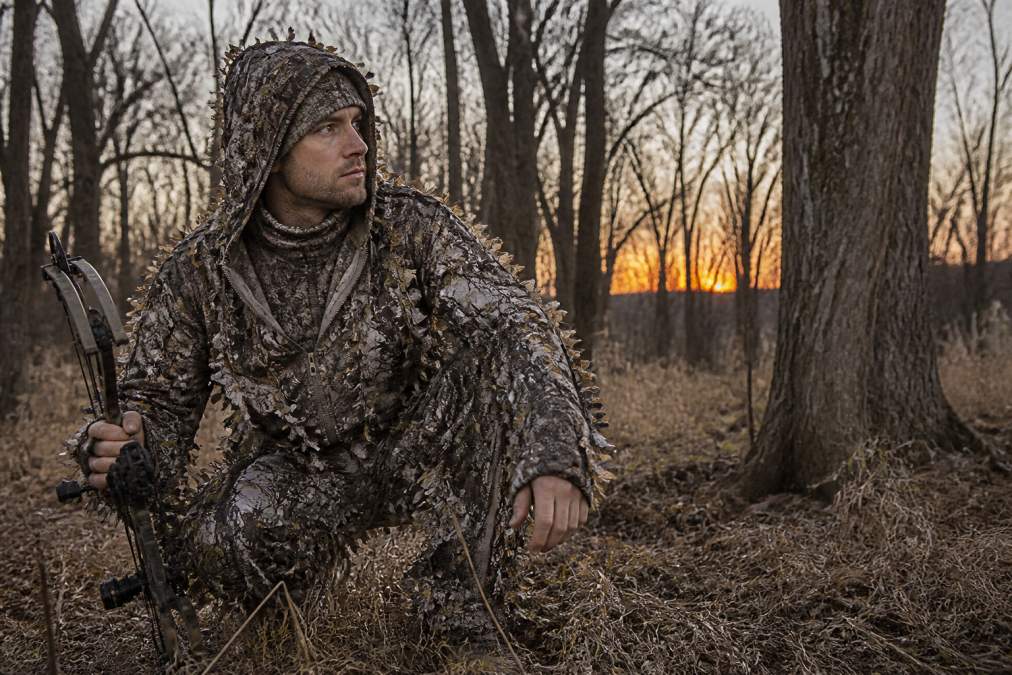 Hunter in full layered outfit crouching in wooded field at dawn, wearing moisture-wicking base layer visible at neck, insulating mid-layer under arms, and camouflage waterproof shell jacket, demonstrating complete layering system in action
