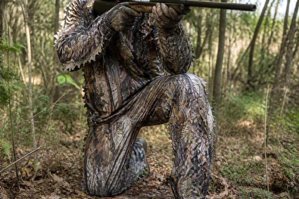 Close-up of hunter crouching in tactical hunting pants with visible articulated knee joints and multi-pocket cargo design, demonstrating mobility and stretch-woven fabric movement through dense brush