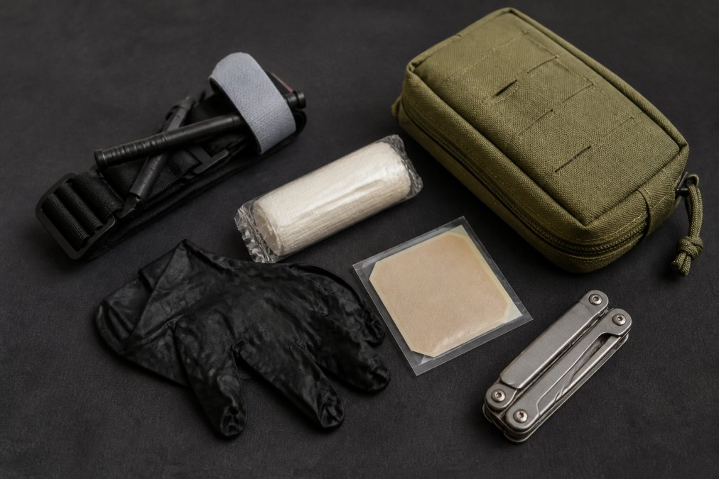 Flat-lay photograph of a compact EDC trauma kit arranged on a dark matte surface, including a CAT tourniquet, rolled hemostatic gauze, a pair of nitrile gloves, a chest seal, and a small olive-drab pouch, with a folded multi-tool beside the kit