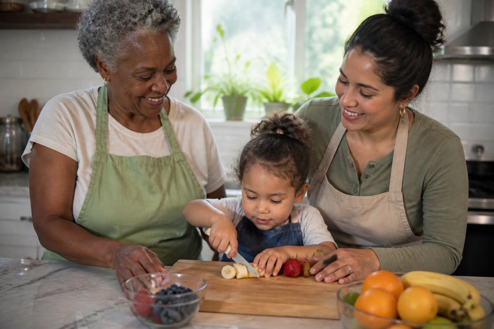 A diverse family helping toddler chefs cut soft fruit with a nylon knife on a wooden board in a bright kitchen.