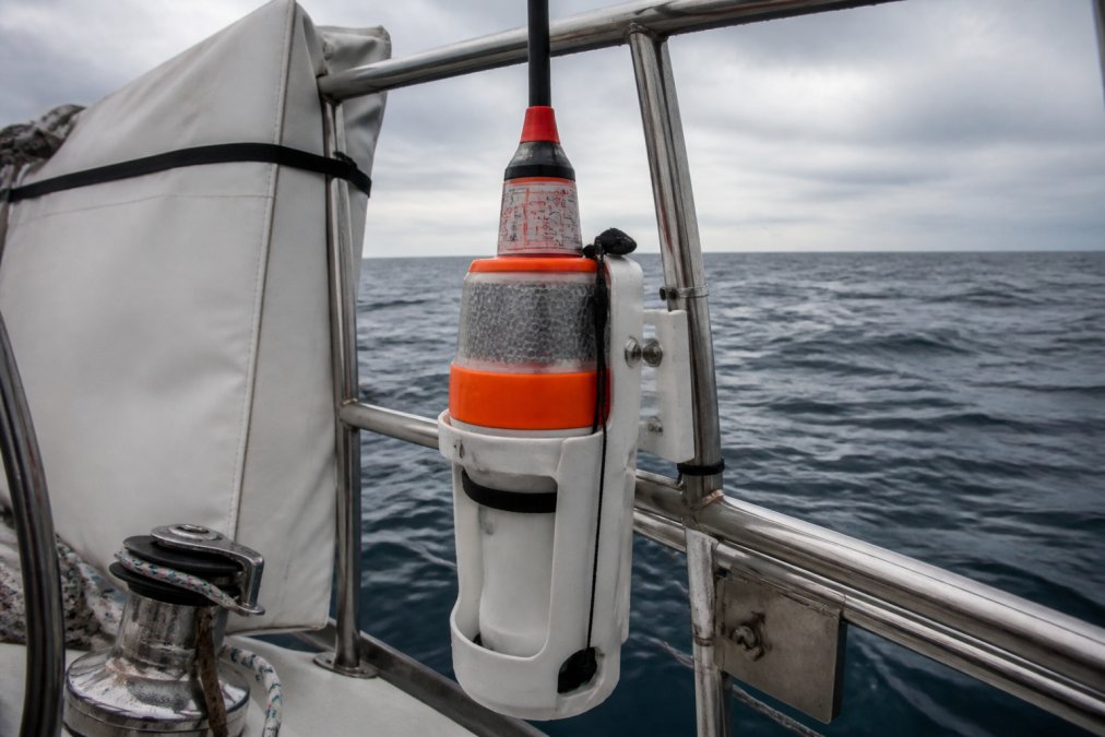 A white and orange EPIRB device mounted securely in its release bracket on the stern rail of a sailboat, the ocean visible in the background under overcast skies, the device's registration label clearly visible