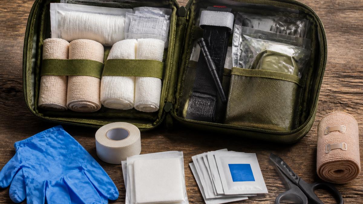 A well-organized open medical kit laid out on a wooden table, showing rolled gauze, bandages, medical tape, non-latex gloves, antiseptic wipes, scissors, and a tourniquet in labeled compartments