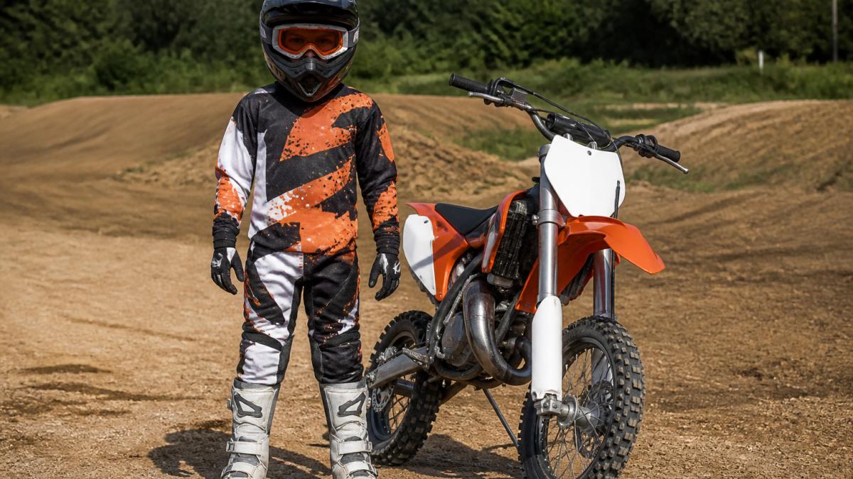 Young beginner motocross rider fully geared up in helmet, goggles, jersey, pants, gloves, and boots standing next to dirt bike in outdoor riding environment with natural daylight