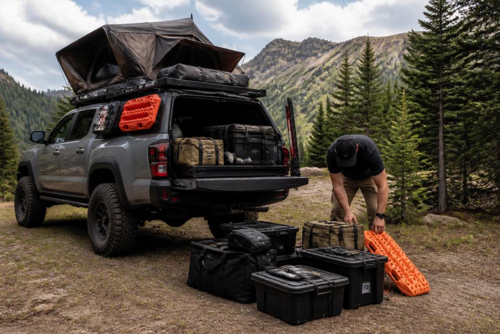 A fully equipped overland truck parked on a scenic mountain trail with visible recovery boards, all-terrain tires, rooftop tent, and organized cargo storage systems mounted on the vehicle