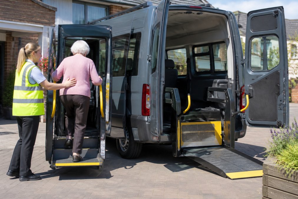 A care home support worker in a high-visibility vest assisting an elderly passenger using a retractable step and handrail to board a modern accessible minibus, with a deployed wheelchair ramp visible at the rear doors, bright daylight, residential care home setting