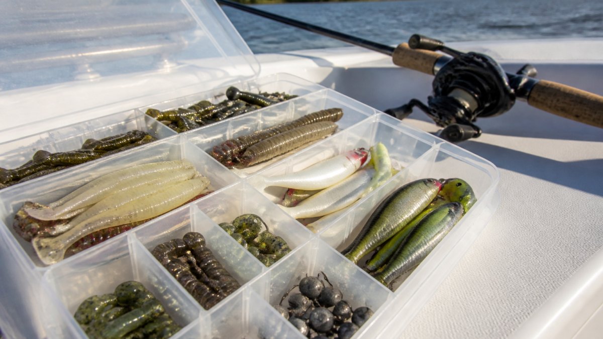 Close-up of an open tackle box on a boat deck showing soft plastic lures, jig heads, and topwater plugs with a fishing rod in the background under natural sunlight