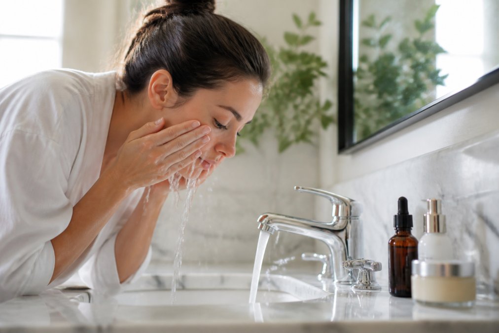 Professional woman at bathroom sink performing morning skincare routine, applying cleanser with warm water, with serum and moisturizer bottles visible on marble counter in natural light
