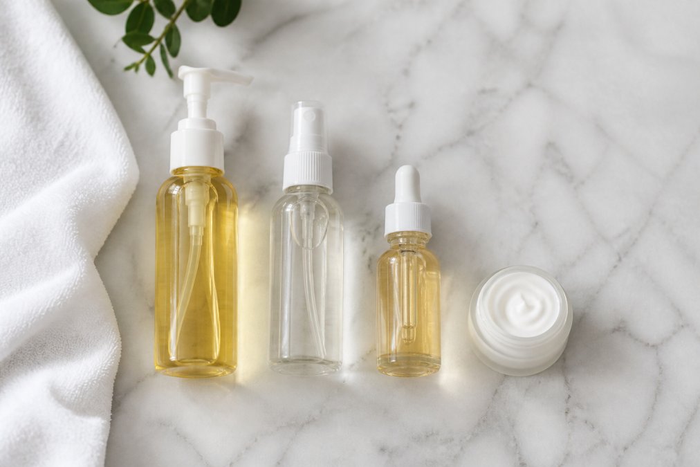 A flat-lay photo of a complete skincare routine on a clean white marble surface, featuring a cleansing oil bottle, a toner in a glass spray bottle, a serum with a dropper, and a small cream jar, arranged in the order they are applied