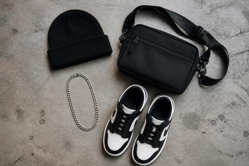 Black beanie, crossbody bag, silver chain, and sneakers arranged as essential streetwear accessories on concrete.