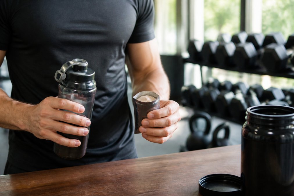 Close-up of athlete's hands holding a water bottle and supplement powder packet, modern gym environment with dumbbells and kettlebells visible in blurred background, bright natural lighting from gym windows