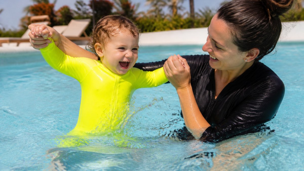 Young toddler in bright high-visibility neon swimsuit holding onto parent's hands while standing in shallow pool water, both smiling with water splashing around them, sunny outdoor pool setting