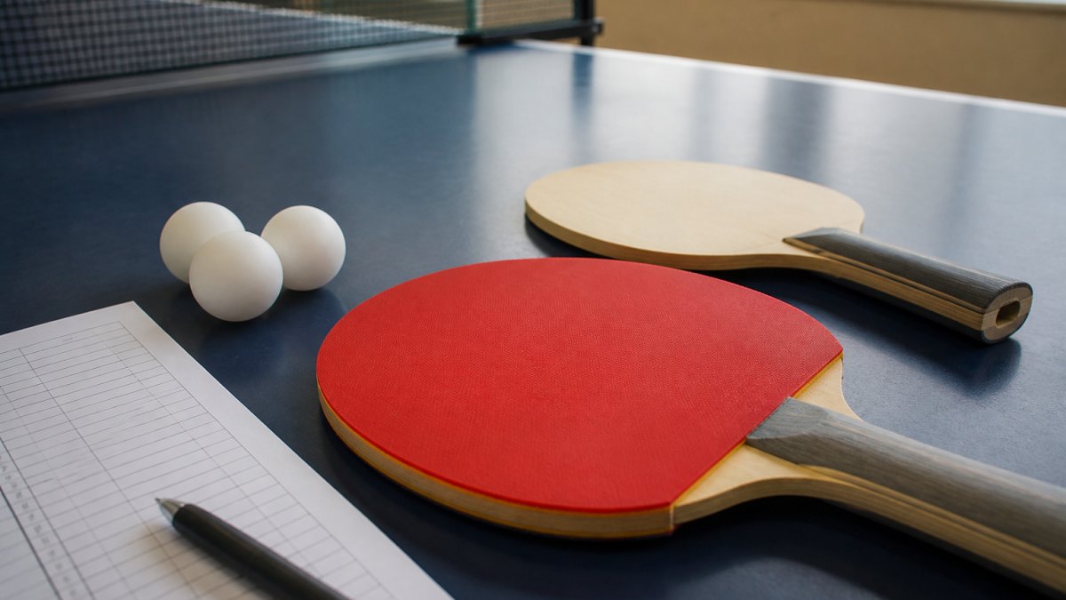 Close-up of competitive table tennis paddle with blade and rubber laid flat on a table next to regulation 40mm balls and a tournament score sheet, natural window lighting