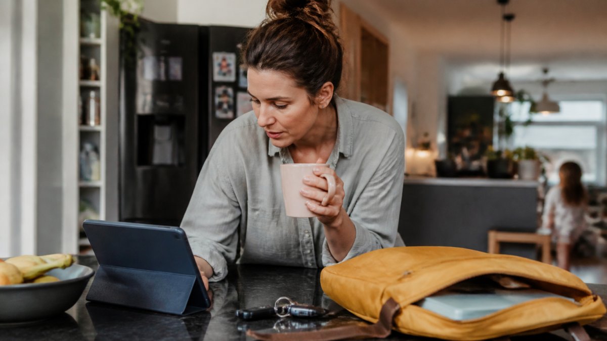 A busy parent checking a smart display on a kitchen counter showing the family calendar, with a child's school bag and keys nearby in a warm, lived-in kitchen