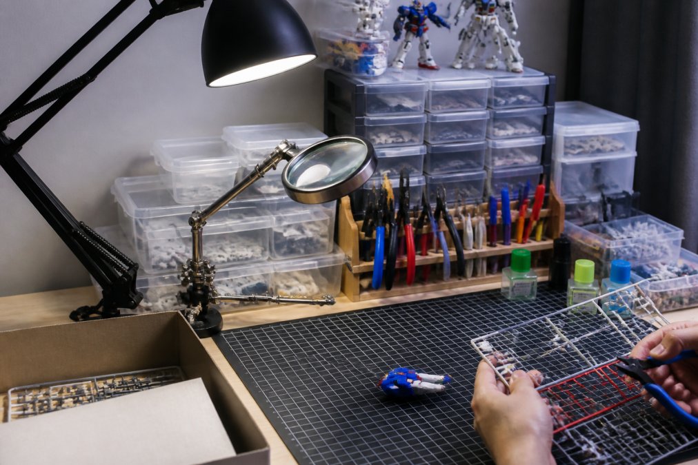 A well-organized hobby workbench with adjustable desk lamp positioned to the side, magnifying glass mounted on an articulating arm, self-healing cutting mat in the center, tool organizer holding nippers and hobby knives, and plastic storage containers holding various model parts and supplies in the background