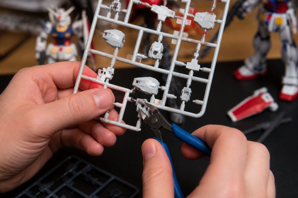 Close-up of hands gripping side cutters positioned against a plastic sprue from a Gundam model kit, cutting a piece cleanly from the runner with proper technique visible, workspace with model parts visible in soft workshop lighting