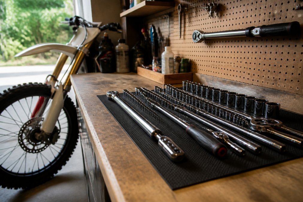 A neat workbench in a home garage with a metric socket set laid out on a mat, a torque wrench hanging on a pegboard, and a dirt bike wheel leaning against the bench in soft natural light