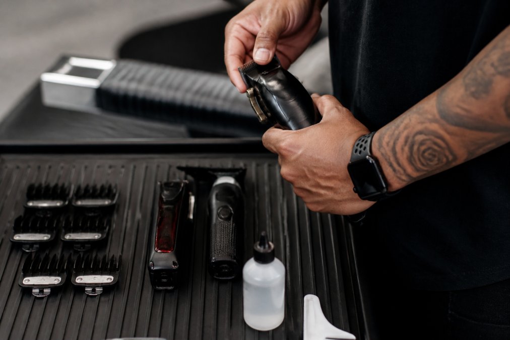 Professional barber's hands holding cordless clippers with guard attachments and blade oil arranged on a station mat in natural lighting