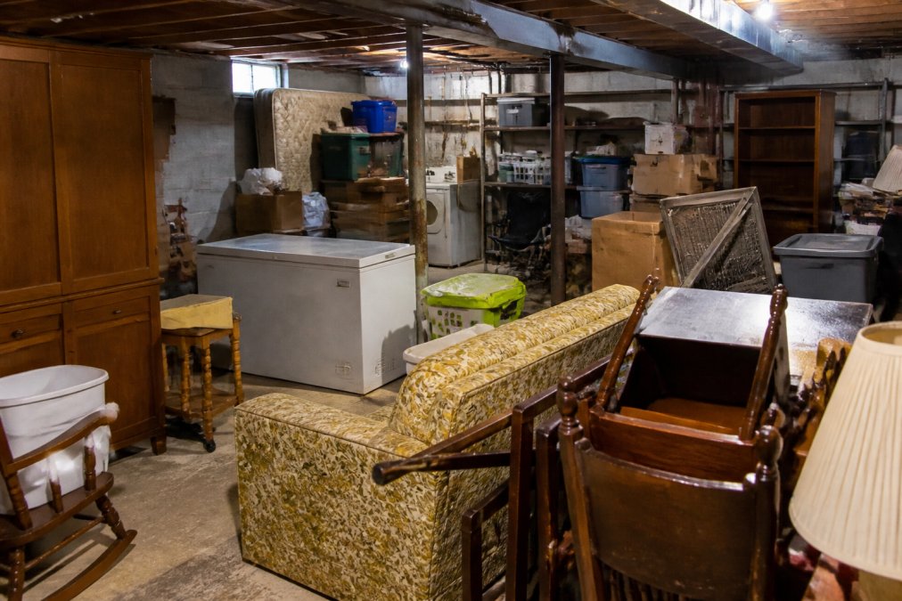 A cluttered basement with furniture, appliances, boxes, and miscellaneous items stacked throughout the space, showing the scope of typical estate cleanup work