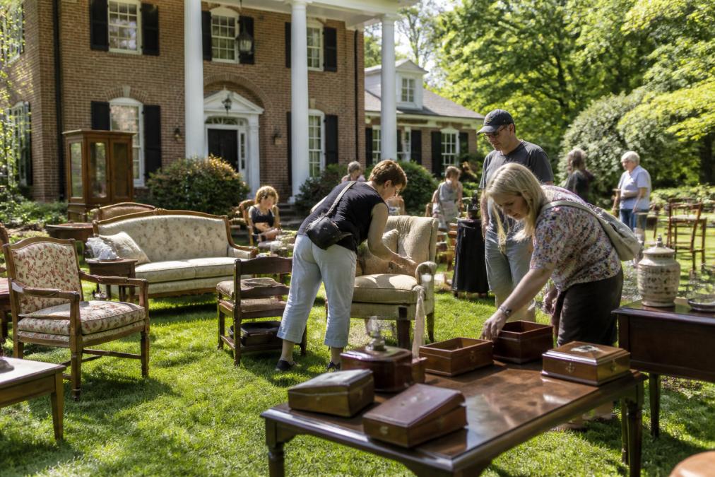 Professional estate sale in progress with organized furniture and household items displayed on the lawn, potential buyers browsing items and examining price tags, morning sunlight across the property