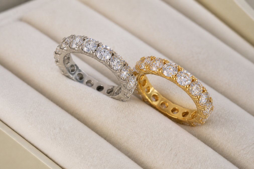 Close-up macro shot of a platinum and 18ct yellow gold eternity ring placed side by side on a white velvet jewellery display, diamonds catching warm studio light to reveal fire and scintillation across the facets