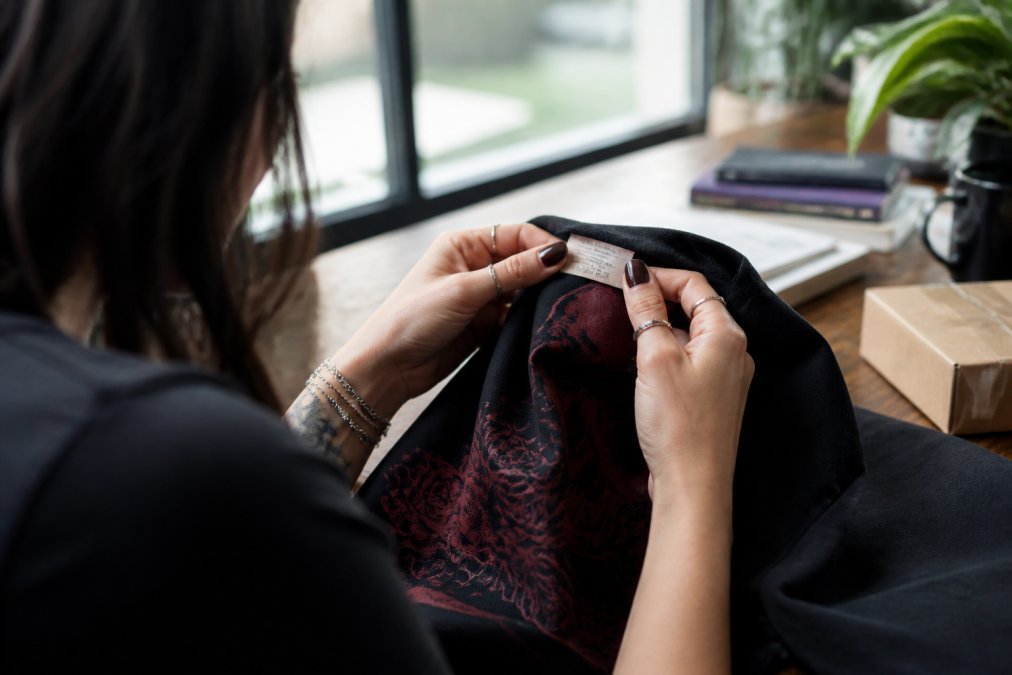 Close-up of hands examining a dark romance-themed graphic tee, holding the fabric to check quality and reading the care label with natural window lighting