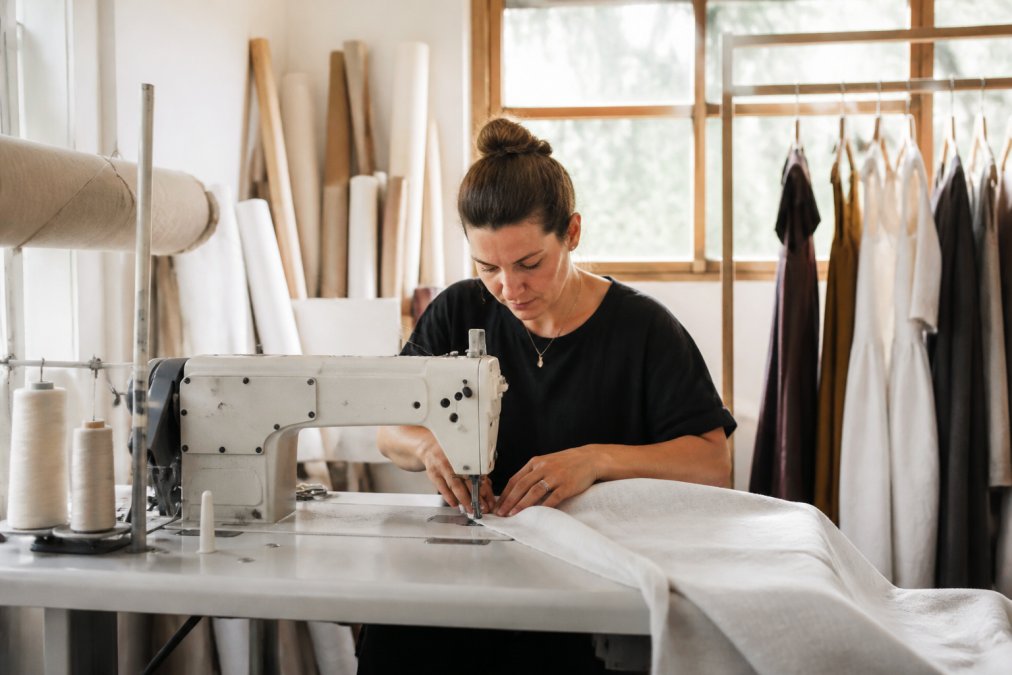 A small-batch garment studio with natural light, a maker sewing organic linen fabric at an industrial machine, rolls of fabric and pattern paper nearby, finished dresses hanging on a rack in the background