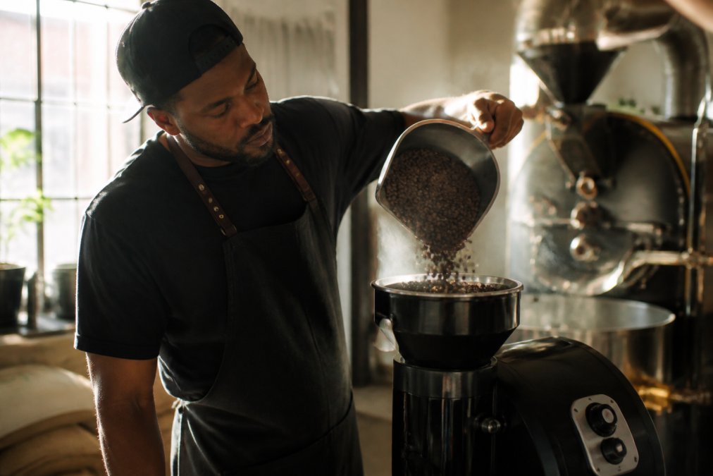 Barista's hands carefully measuring and pouring freshly roasted coffee beans into a grinder in a bright roasting facility, steam rising from the beans with natural window lighting