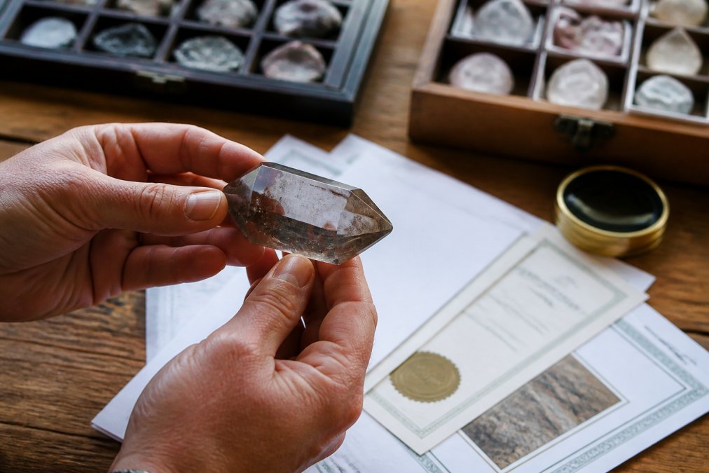 Close-up of a collector's hands examining a polished crystal specimen under natural light, with sourcing documentation and certification papers visible on a wooden table