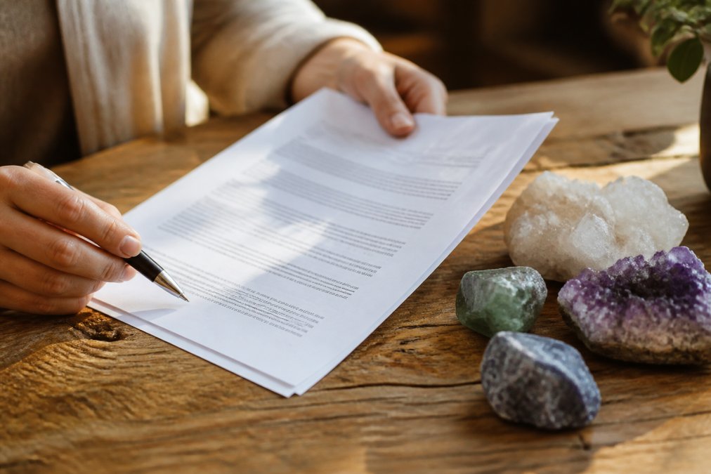 Close-up of hands holding a printed document with a pen, next to a small pile of raw crystals on a wooden table in warm natural light
