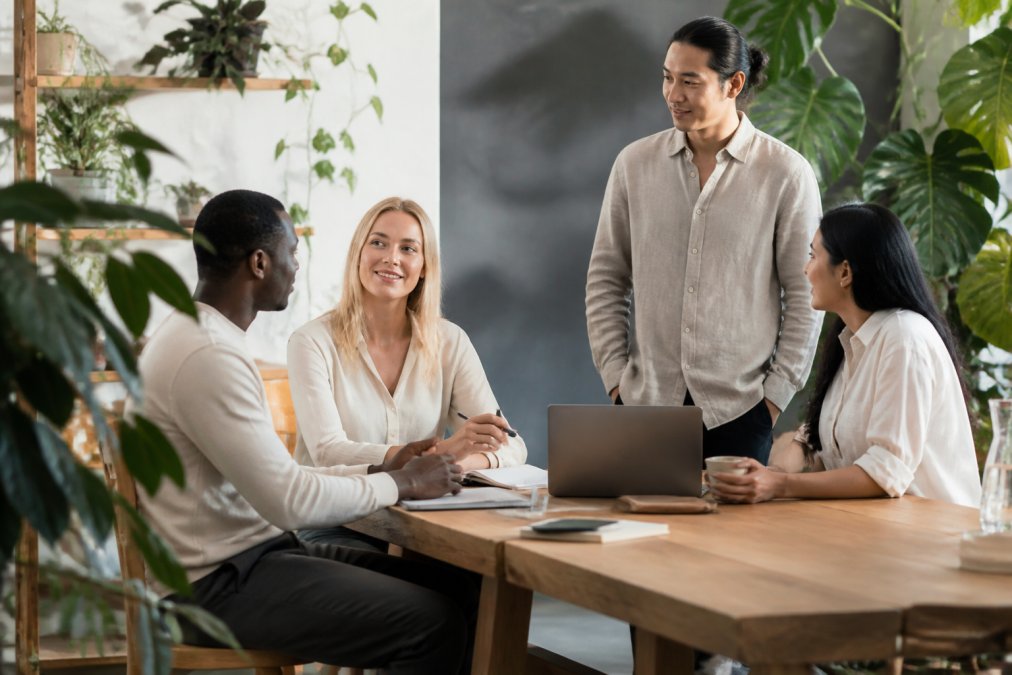 A diverse group of professionals in a bright, modern office, wearing minimalist clothing in natural fabrics like organic cotton and linen, collaborating around a wooden table with laptops and notebooks