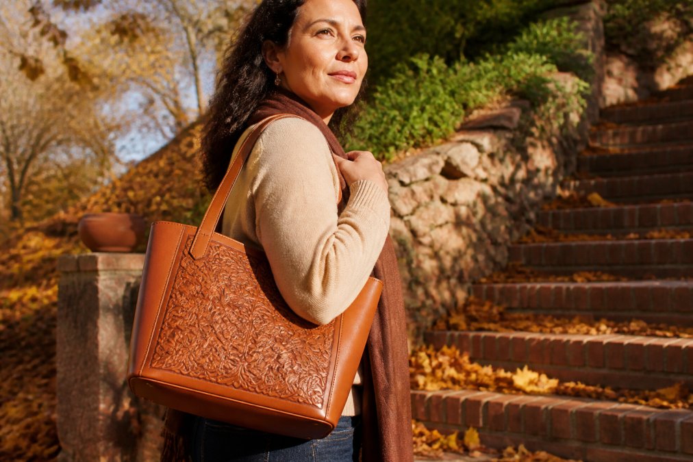 Woman in her 40s wearing a cognac-toned hand-tooled vegan leather tote bag over her shoulder, standing outdoors in autumn setting with warm lighting, showing the bag's detailed craftsmanship and how it pairs with classic fall wardrobe pieces