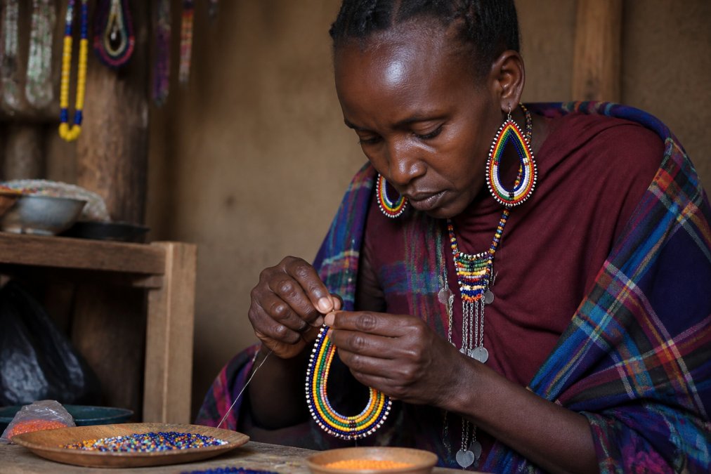 Skilled artisan in an East African workshop carefully hand-crafting a beaded necklace with intense concentration, showing detailed handiwork with traditional beads and wire, natural workshop lighting illuminating the intricate work
