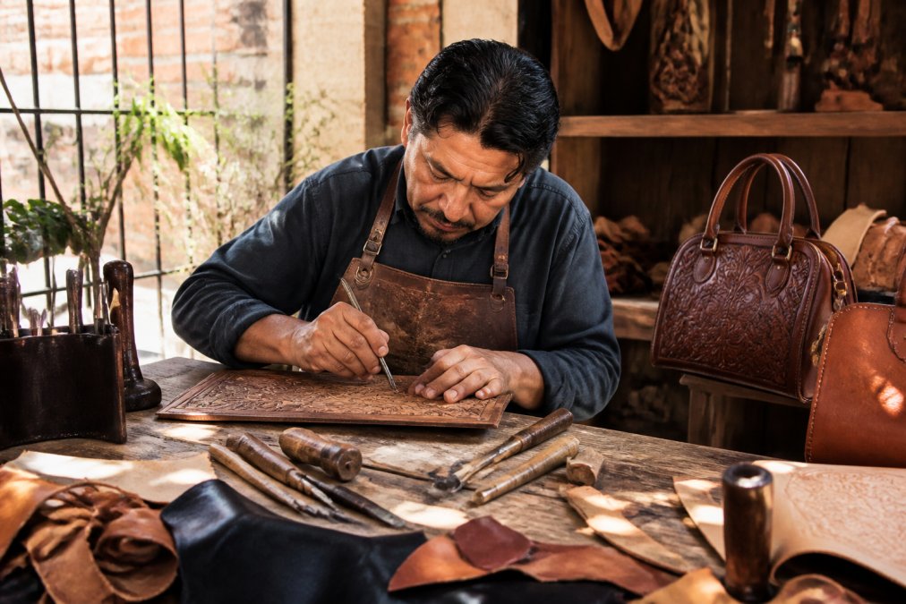 Artisan craftsperson sitting at a wooden workbench in natural sunlight, hand-tooling intricate leather designs with traditional metal tools, focused on detailed work with leather scraps and finished pieces visible