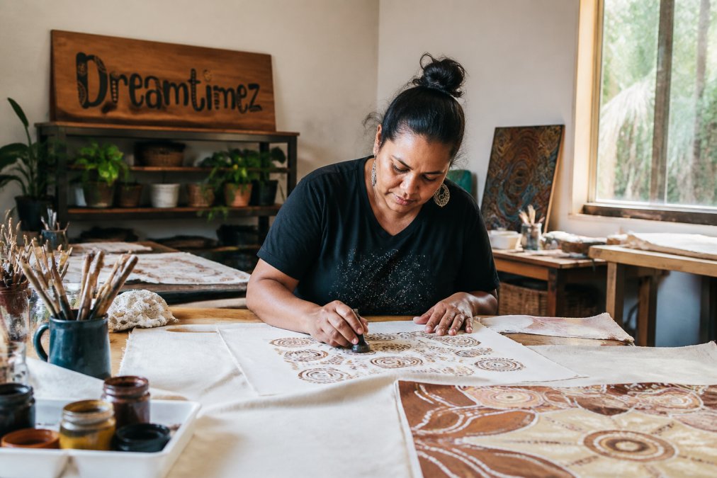 An Indigenous artist working in a studio setting, creating or hand-printing designs on fabric, with natural light and art materials visible