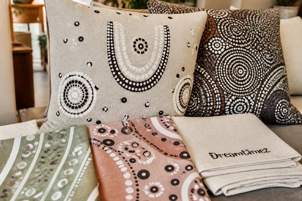 Close-up of Indigenous-designed cushion covers and tea towels displayed together, showing traditional dot motifs and contemporary colour palettes on natural linen fabric in soft natural lighting