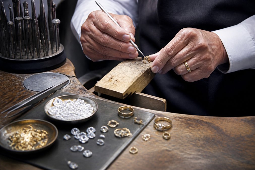 Close-up of a master jeweler's hands working with precision tools at a polished workbench, surrounded by lab-grown diamonds, recycled gold, and handcrafted components under bright studio lighting