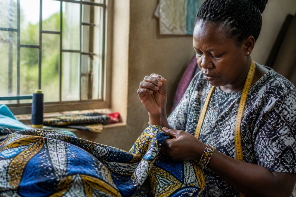 Female artisan hands working intricately with fabric and needle, handcrafting detailed textile work in natural daylight from a workshop window, showing fine needlework detail and vibrant patterned cloth