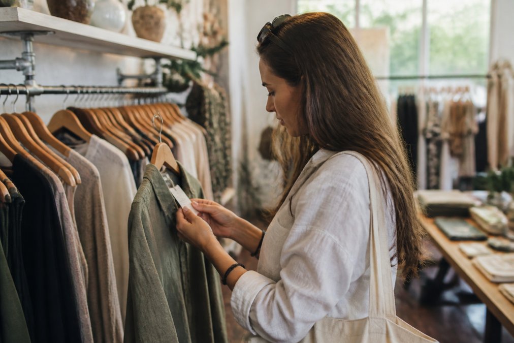 A shopper in a clothing store examining a garment tag closely, with racks of clothes in the background, natural lighting
