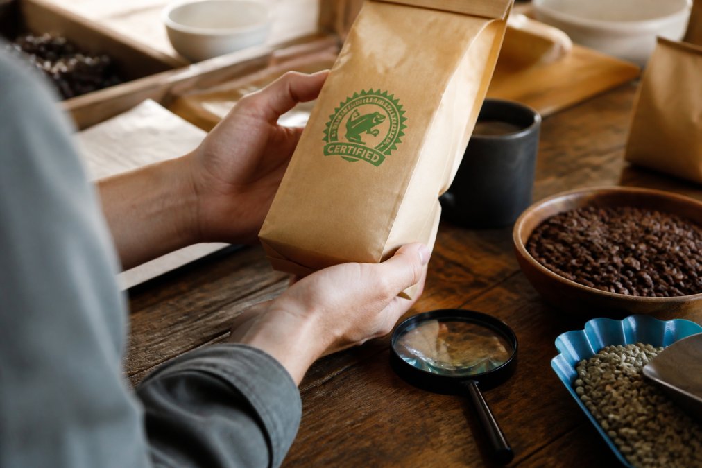 Close-up of a person's hands holding a coffee bag with a certification label, a magnifying glass resting nearby on a wooden counter, warm natural light