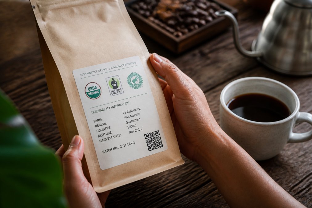Close-up of hands holding a coffee bag showing certification labels and traceability information, with a cup of brewed specialty coffee nearby