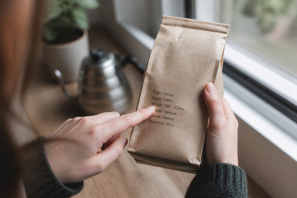 Close-up of a person's hands holding a kraft paper coffee bag, index finger pointing at the printed origin, altitude, and processing details on the label, soft daylight from a window
