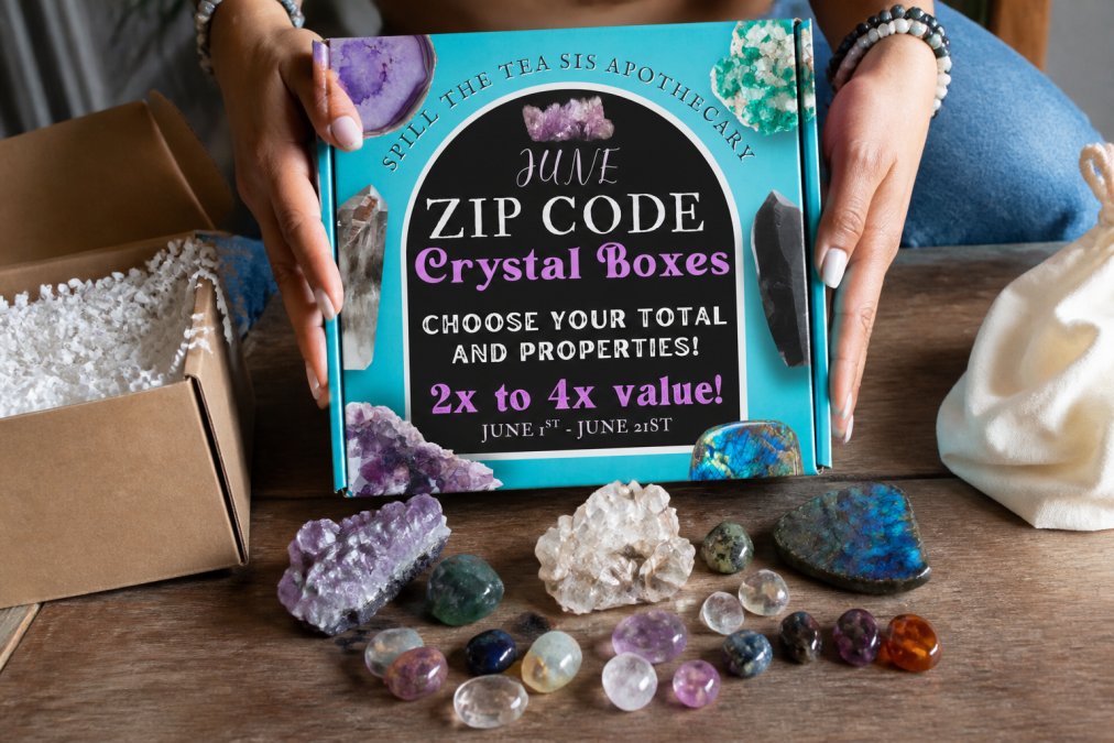 Hands opening a mystery crystal box, revealing tumbled stones, raw gemstones, and crystal clusters spilling out onto a wooden surface with soft natural lighting