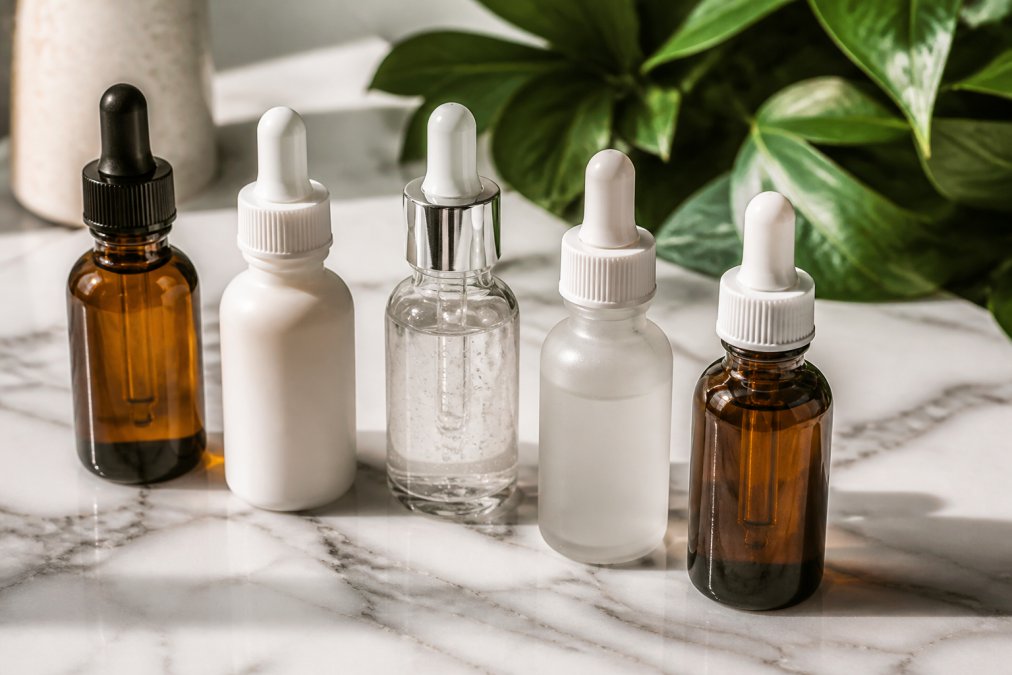 A flat lay of five EVERGRENE serum bottles in amber and white arranged neatly on a white marble surface, soft natural light, green botanical leaves blurred in the background