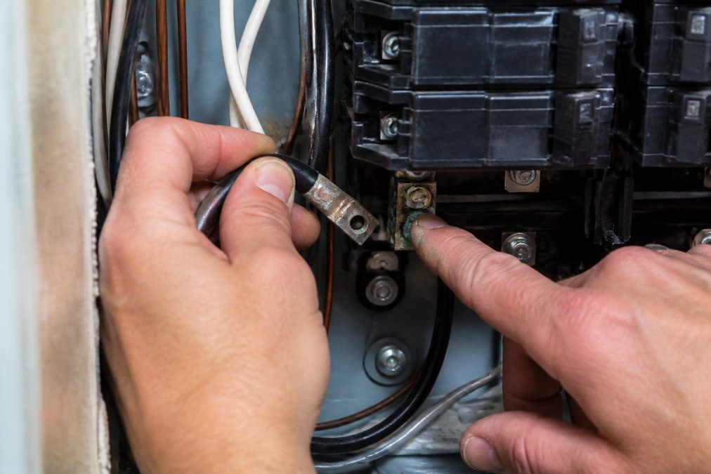 Professional electrician's hands inspecting a loose terminal connection at an electrical panel, showing corrosion and discoloration on the contact point under bright panel lighting