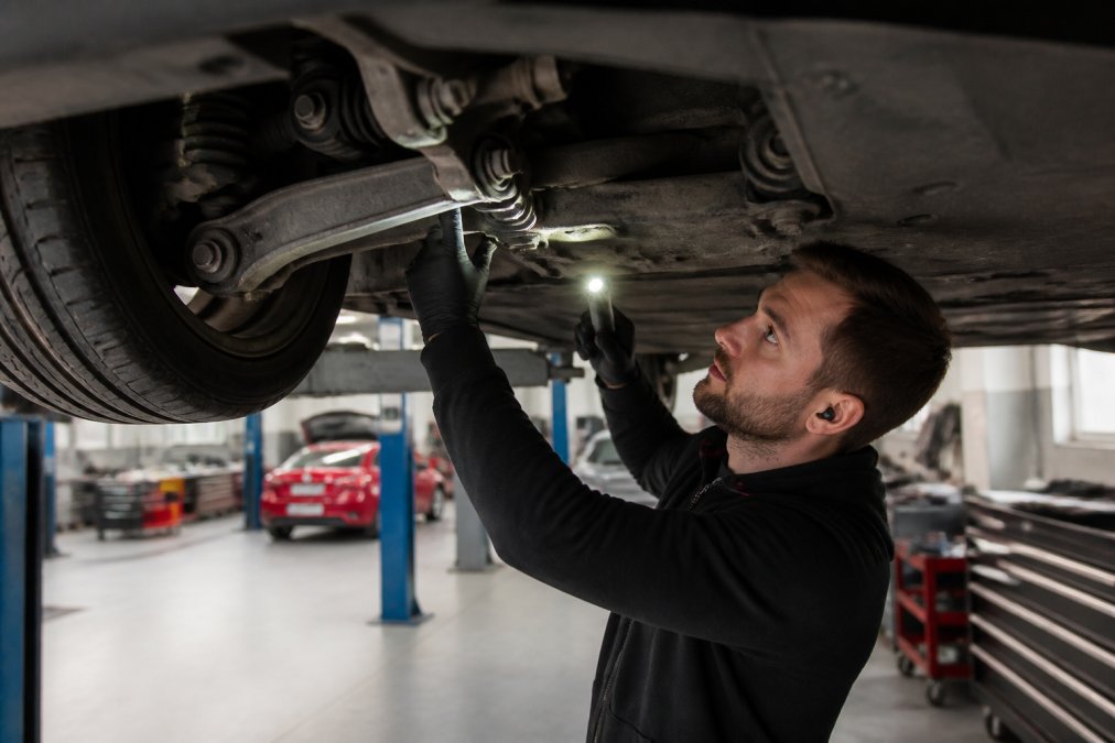 Professional technician performing visual suspension inspection under electric vehicle on lift, examining control arms and bushings with torch light in modern workshop