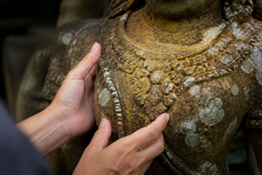 Close-up of an antique stone statue surface showing natural golden-brown patina, lichen growth patterns, and weathered texture with hands carefully examining the carved details under soft natural light