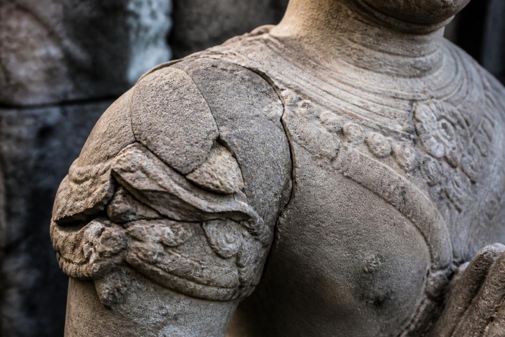 Detailed view of an antique stone statue showing visible hairline cracks running through carved areas, spalling edges with chunks missing, and delamination along layered surfaces, highlighted by angled natural light