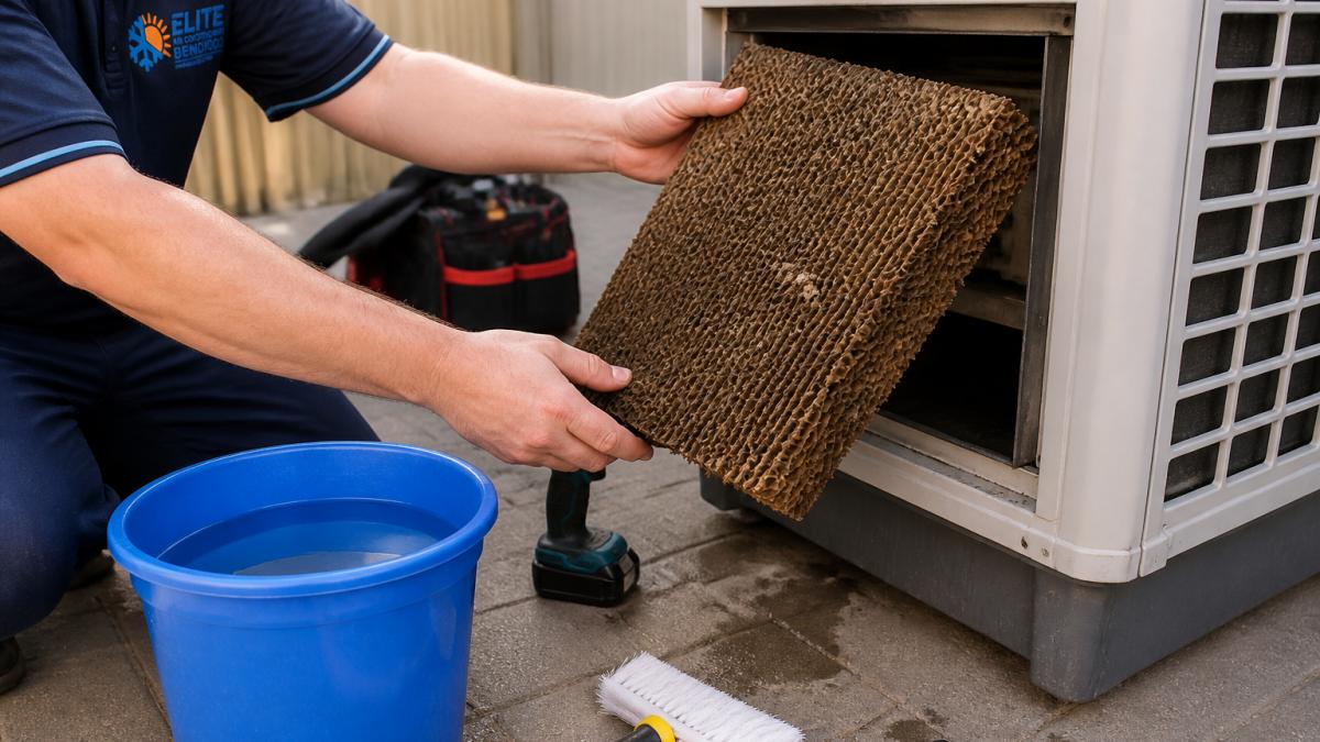 Close-up of hands removing a dusty evaporative cooler pad from the unit's side panel, with a bucket of water and cleaning brush nearby on a paved outdoor area