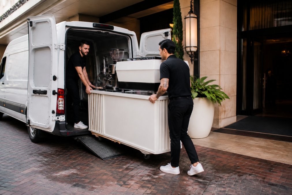 Professional barista team unloading a mobile coffee cart from a van at an upscale event venue entrance, carefully maneuvering the equipment down a ramp while a venue coordinator gestures directions