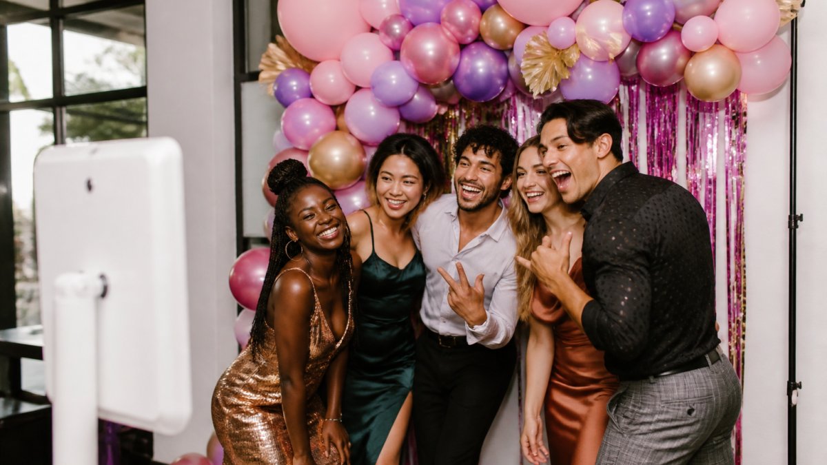A group of friends in colorful party outfits posing and laughing in front of an iPad photo booth at a birthday party, with a balloon arch and streamers in the background